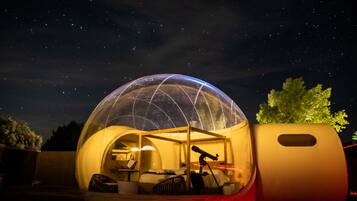 Una estructura en forma de cúpula transparente con una cama y un telescopio en su interior, frente a un cielo nocturno.