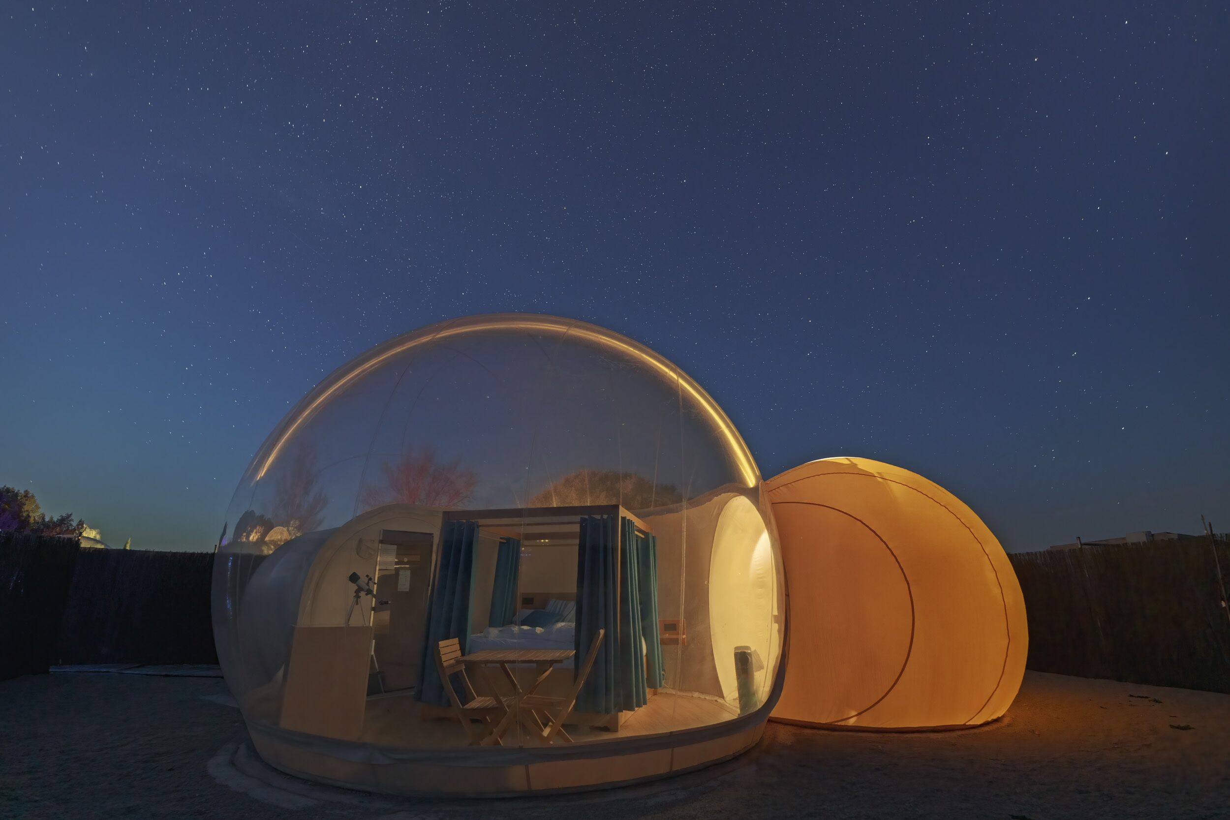 A transparent dome structure with an interior seating area and a bed, set against a night sky with stars.