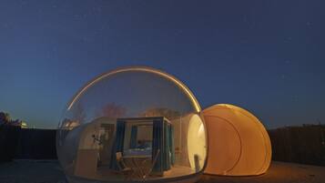 A transparent dome structure with an interior seating area and a bed, set against a night sky with stars.