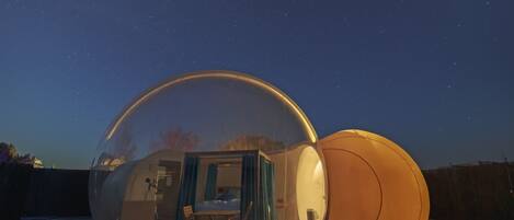 A transparent dome structure with an interior seating area and a bed, set against a night sky with stars.