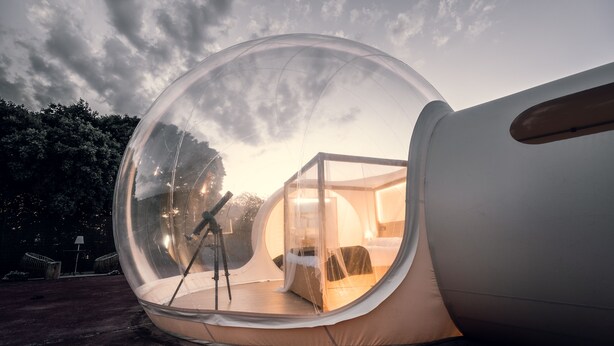 Una estructura de cúpula transparente con una cama y un telescopio en su interior, sobre un fondo de árboles y un cielo nublado.