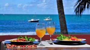 Una mesa con platos de comida y dos vasos de jugo de naranja, frente a una vista del océano con barcos y palmeras.