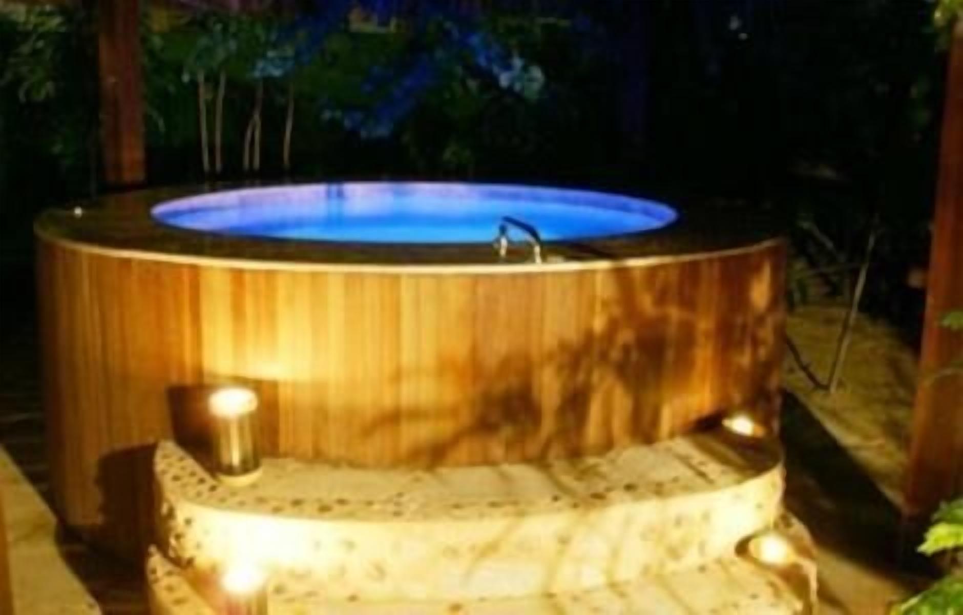 A hot tub with a wooden exterior and a blue interior, surrounded by a wooden deck and two small lamps.