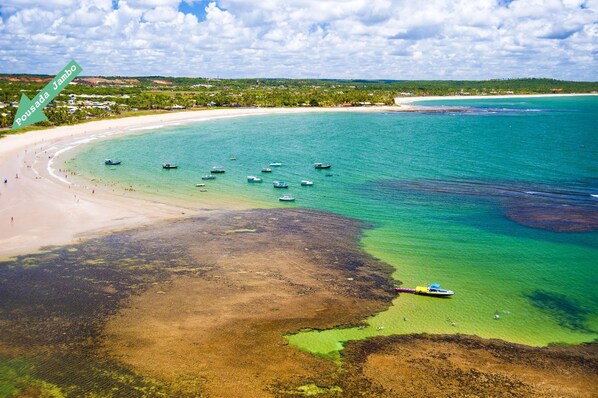 An aerial view of a beach with boats, clear water, and a green arrow pointing to Pousada Jambo. - Pousada Jambo (Camaçari)