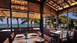 Un área de comedor al aire libre con mesas dispuestas para dos, con vista a una playa con palmeras y el océano.