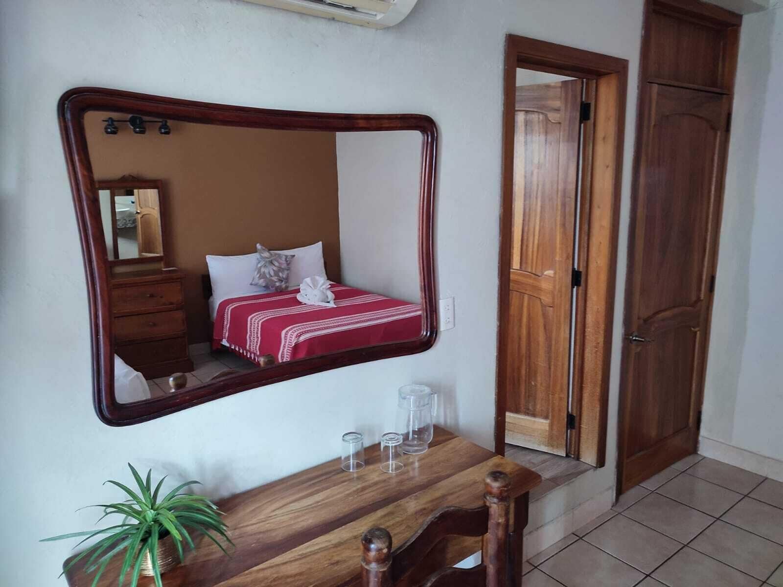 A bedroom with a large mirror, a wooden dresser, a bedside table with glasses, a potted plant, and a wardrobe.