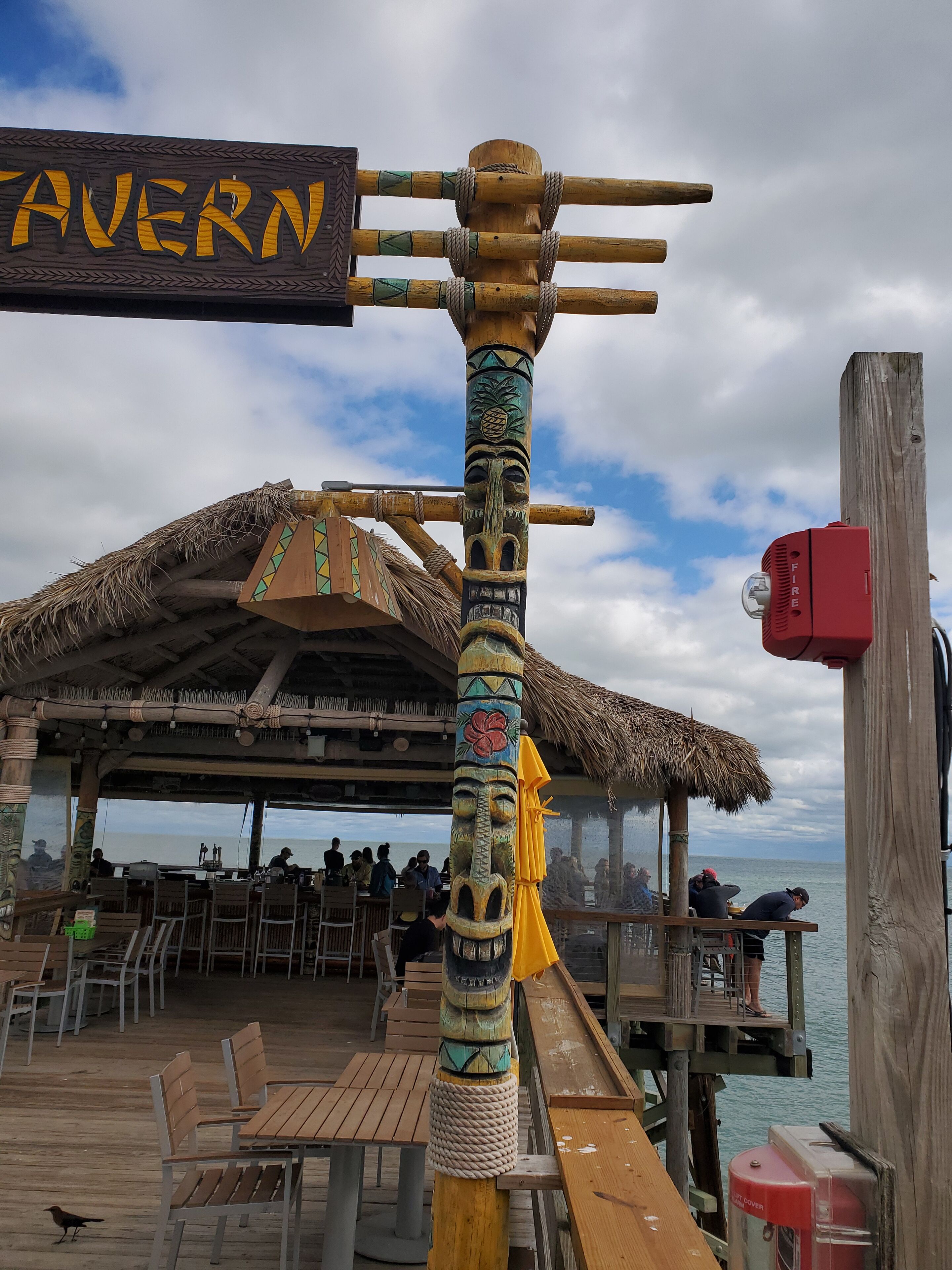 A wooden sign with "TAVERN" written on it, a thatched roof structure, wooden tables and chairs, and a view of the sea.