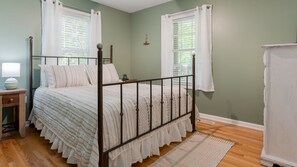 Une chambre à coucher avec un lit à baldaquin, une table de chevet avec une lampe, une commode et une fenêtre ornée de rideaux blancs.