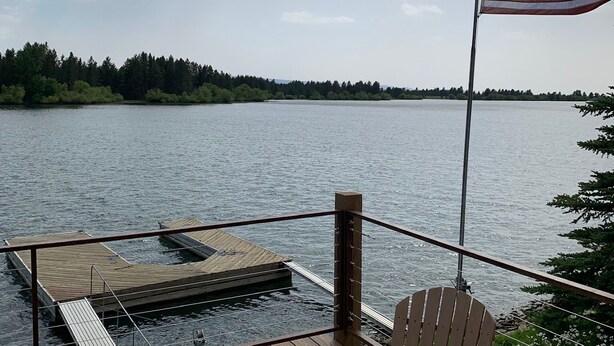 Terraza de madera con un jacuzzi y una silla, con vista a un lago y una bandera estadounidense.