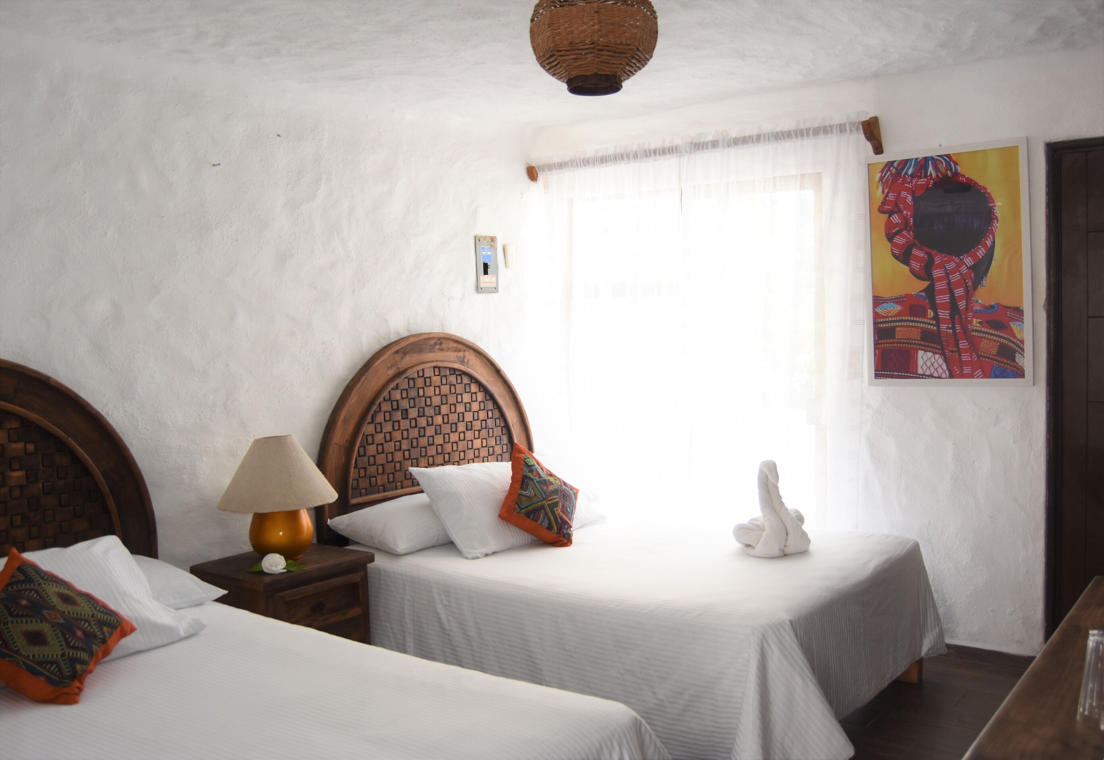 A bedroom with two single beds, a nightstand with a lamp, a decorative wall hanging, and a window with curtains.