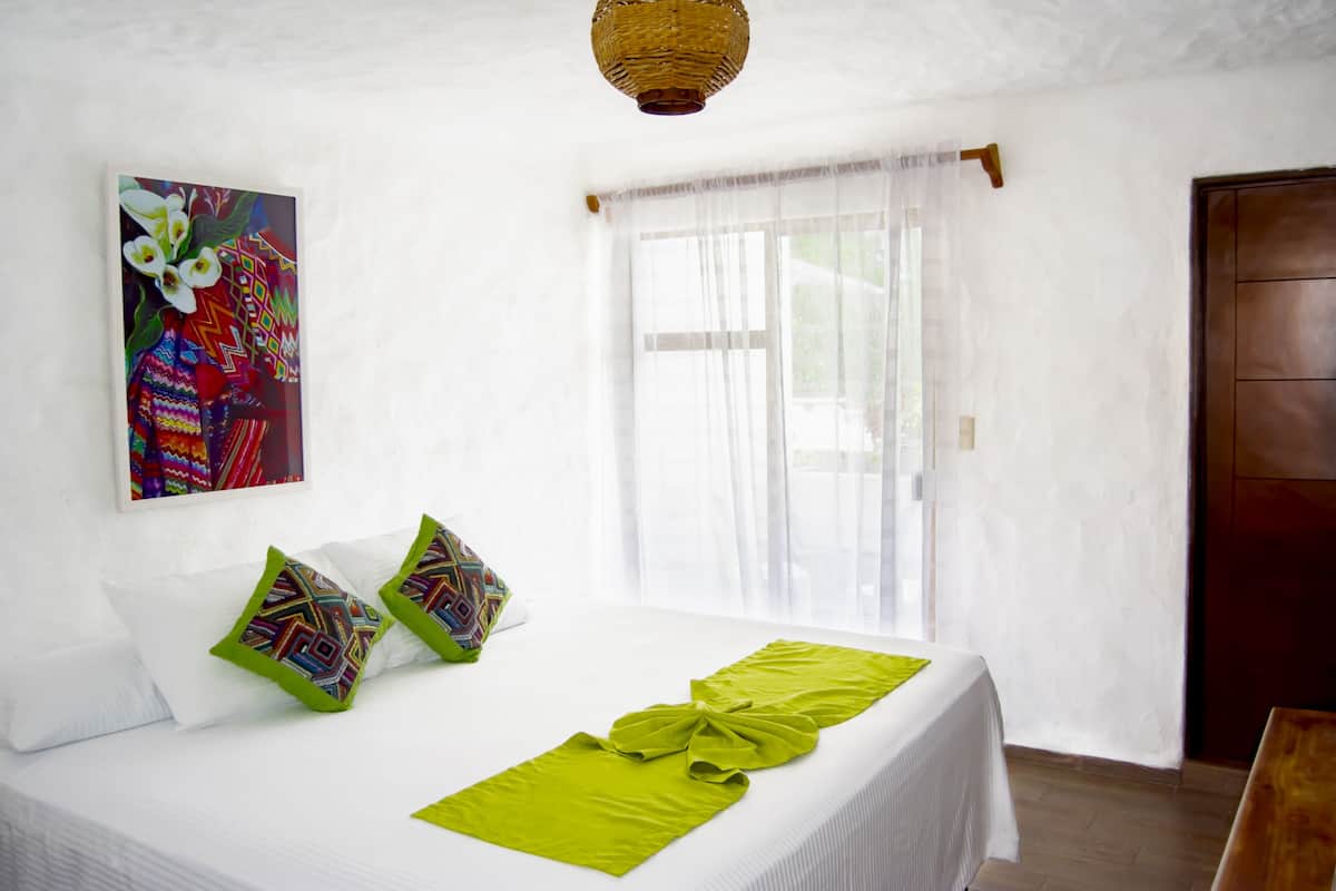 Une chambre d’hôtel avec un lit, une fenêtre ornée de rideaux légers, une porte en bois et un tableau coloré accroché au mur.