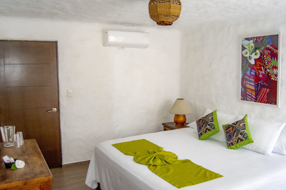 Une chambre d’hôtel avec un lit, une table en bois sur laquelle sont posés des verres, une lampe et un élément de décoration murale aux couleurs vives.