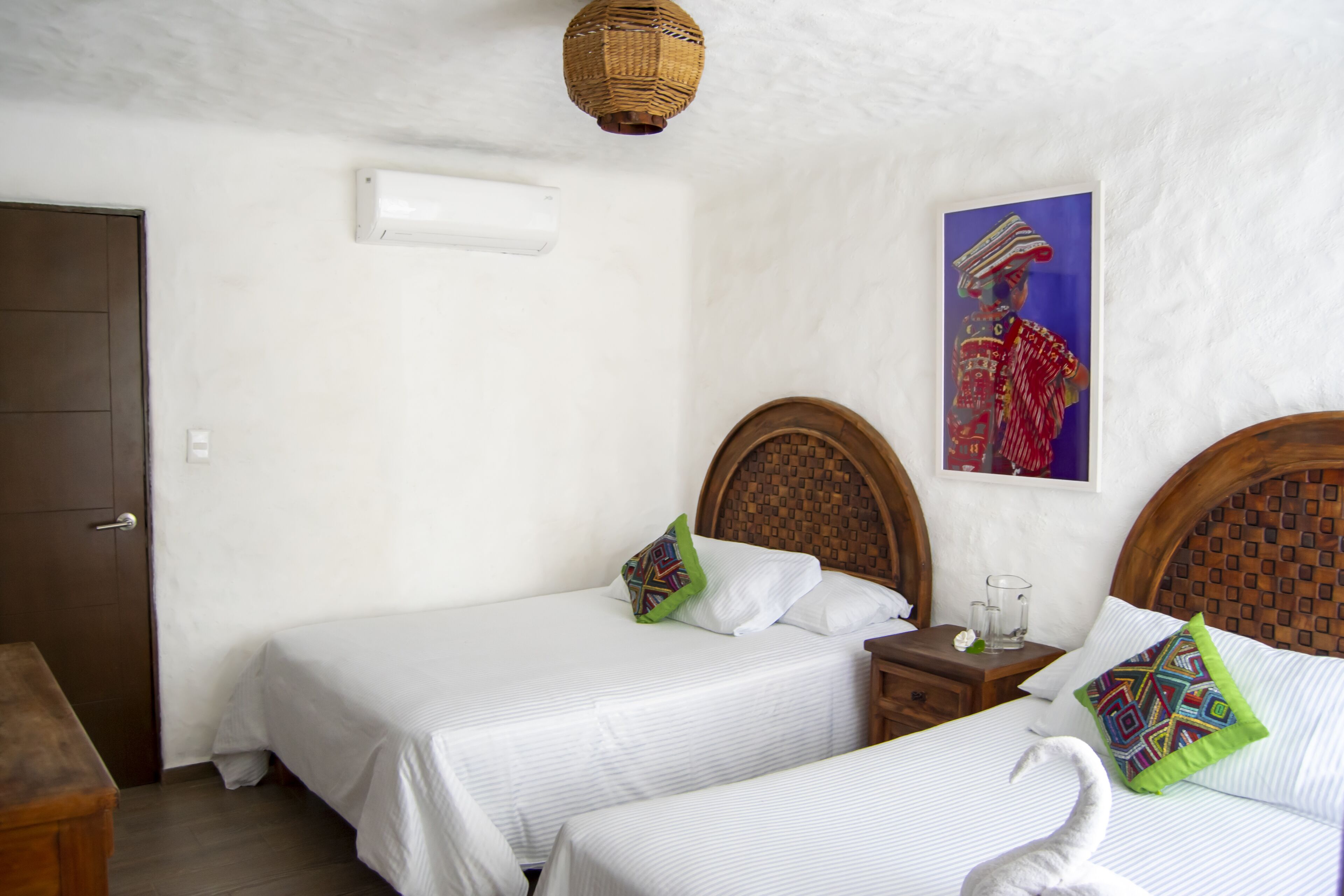 A hotel room with two single beds, a wooden headboard, a small bedside table with a glass, and a wall-mounted air conditioner.