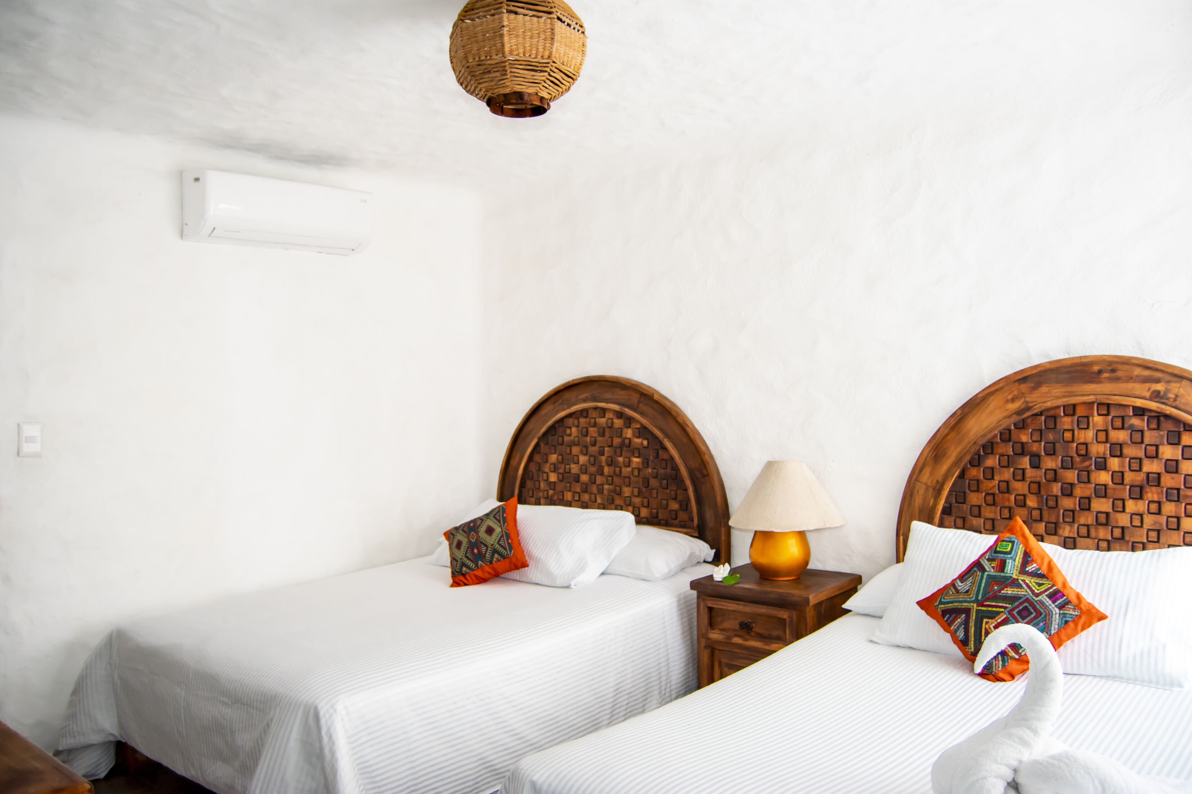 A hotel room with two single beds, a wooden headboard, a bedside table with a lamp, and a wall-mounted air conditioner.