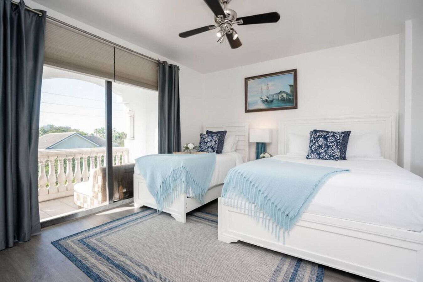 A hotel room with two beds, a ceiling fan, a large window with curtains, and a painting on the wall.
