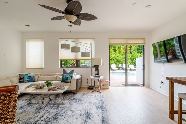 A modern living room with a ceiling fan, a sofa with cushions, a coffee table, and a view of a pool through glass doors.