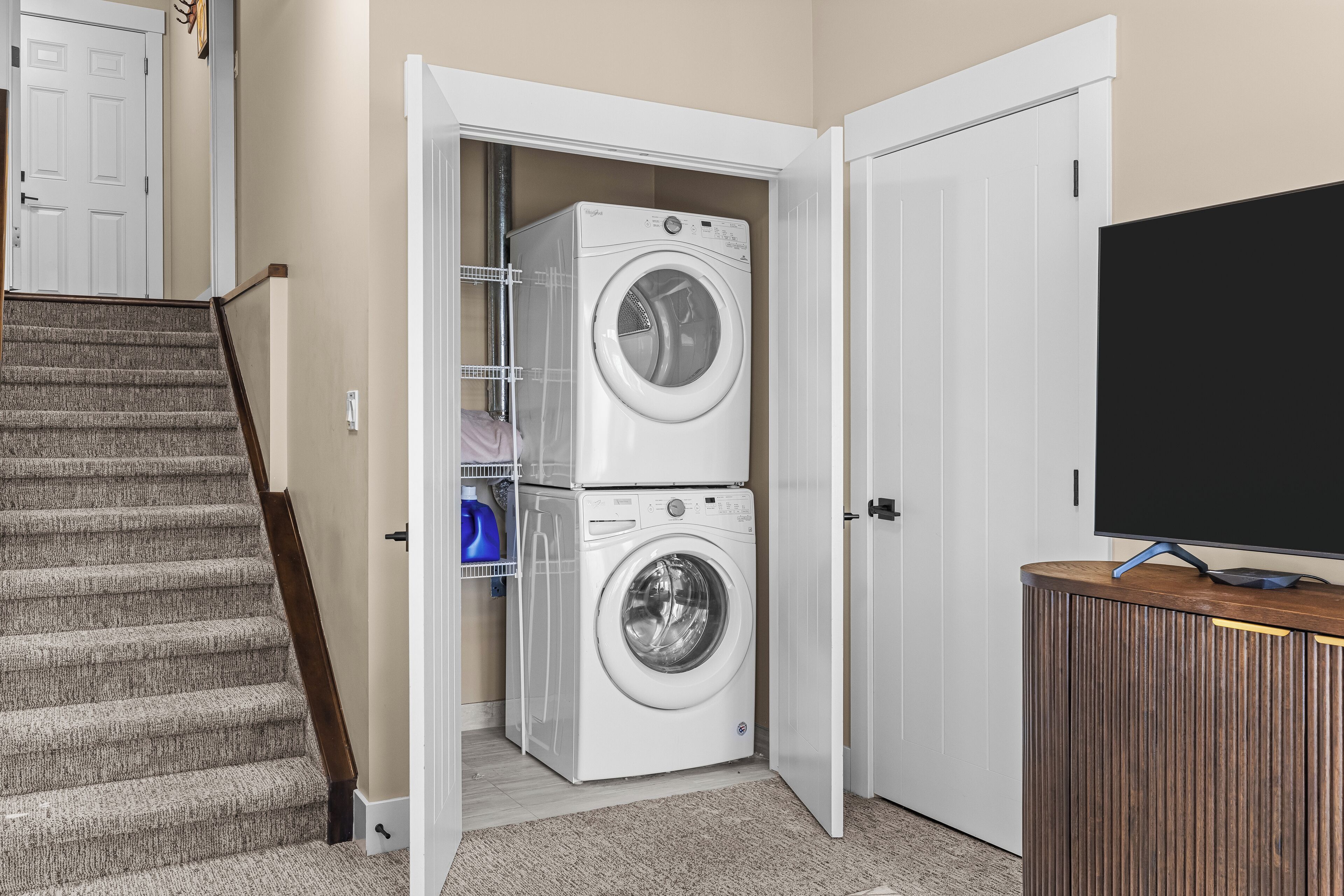 A laundry room with a stacked washer and dryer, a television on a wooden cabinet, and a carpeted staircase.