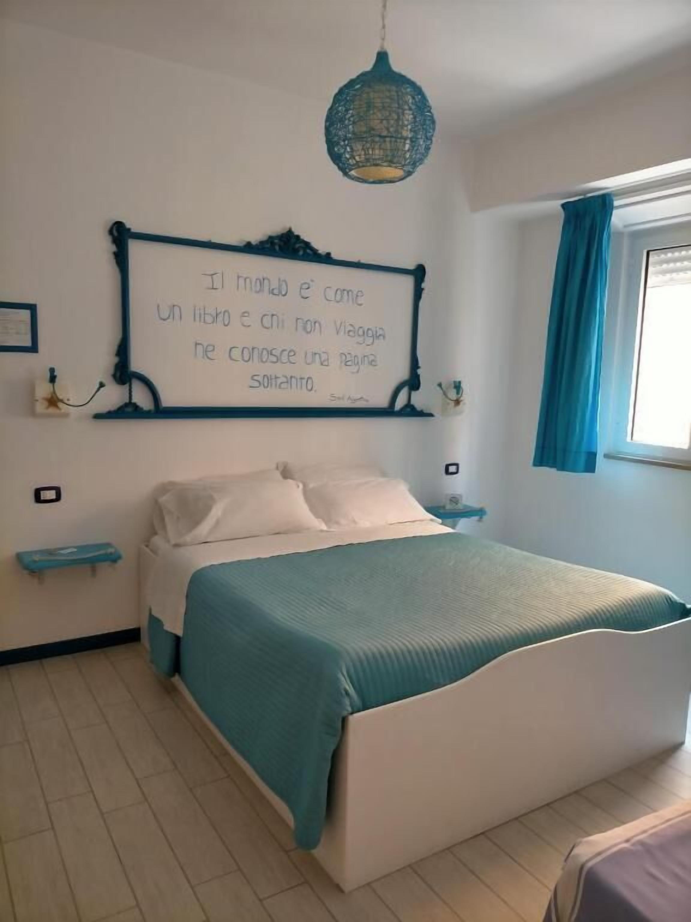 A bedroom with a bed, a nightstand, a wall-mounted shelf, a hanging light fixture, and a window with curtains.