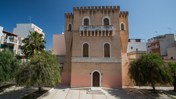 A historic building with a central arched entrance, a balcony with railings, and a tower with battlements. - ControVento Rooms (Margherita di Savoia)