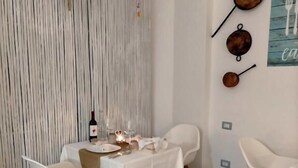A dining area with a table set for two, white chairs, and a decorative wall with wall art and a string of lights. - ControVento Rooms (Margherita di Savoia)