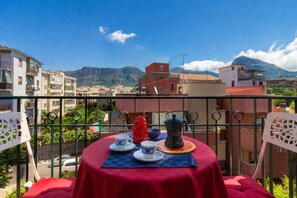 Uma varanda com mesa vermelha posta para dois, com vista para uma paisagem urbana com edifícios e montanhas.