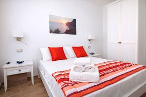 Quarto de hotel com uma cama, duas mesinhas de cabeceira, um criado-mudo, uma luminária na parede, um quadro e um guarda-roupa.