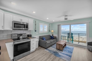 A modern kitchen with a microwave, stove, and oven, a gray sofa, a wooden coffee table, and a view of the beach through a sliding glass door.