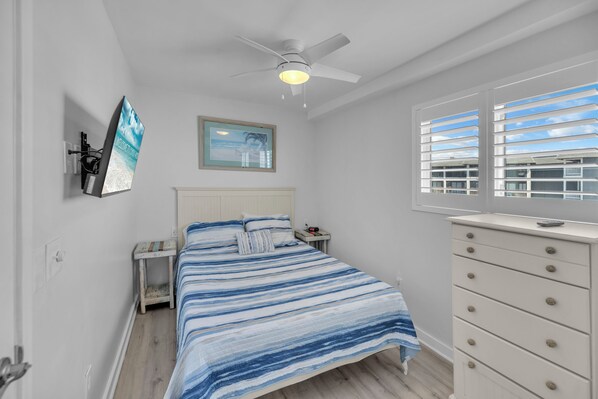 A bedroom with a bed, a nightstand, a dresser, a ceiling fan, a window with blinds, and a wall-mounted TV.