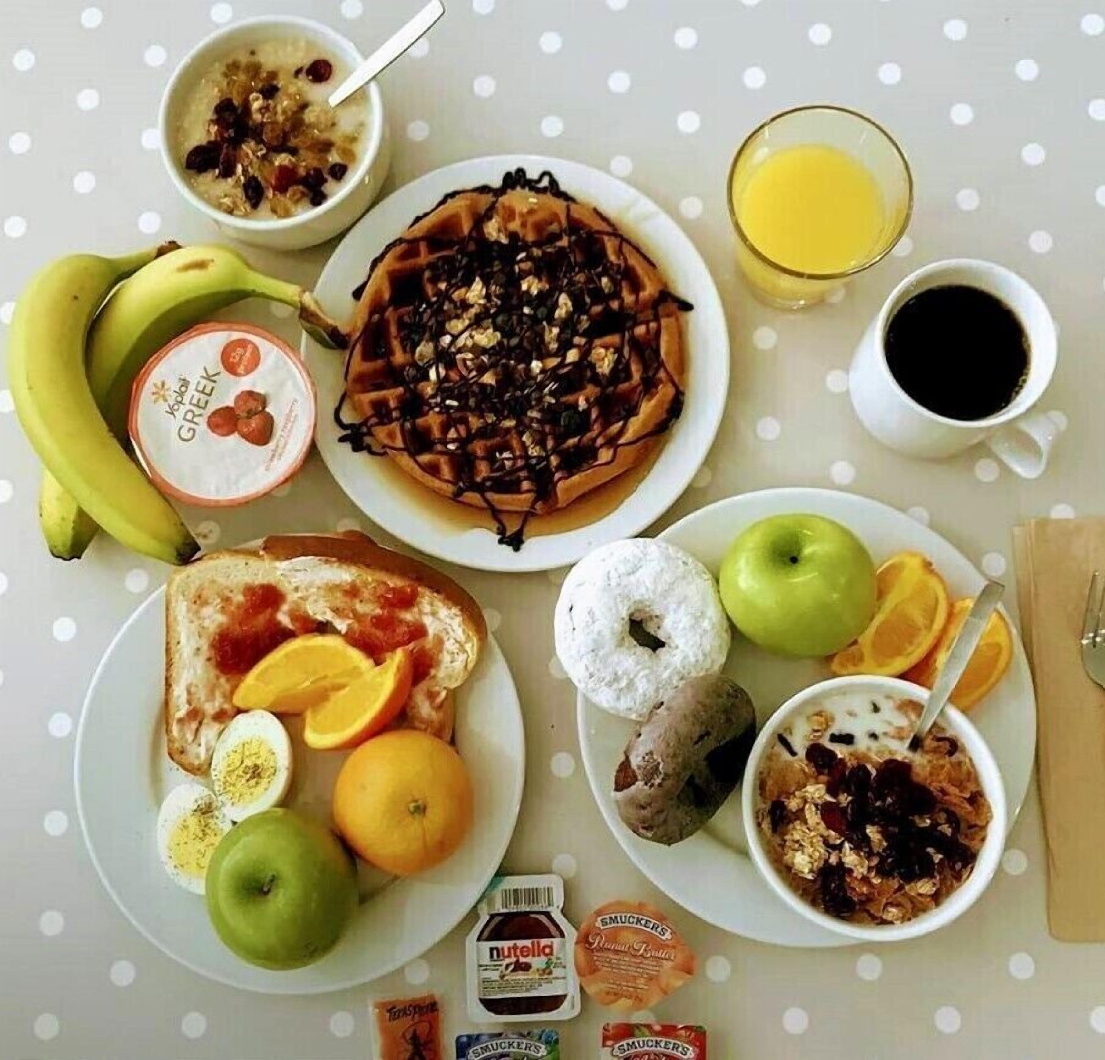 Mesa de café da manhã com diversos alimentos, incluindo waffle, torradas, iogurte, frutas e um copo de suco de laranja.