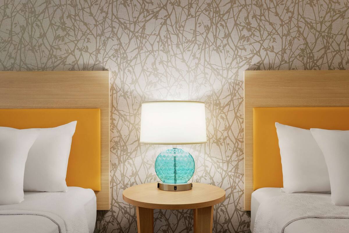 A hotel room with two beds, each with a wooden headboard and white bedding. A round wooden table with a globe lamp is centered between the beds.