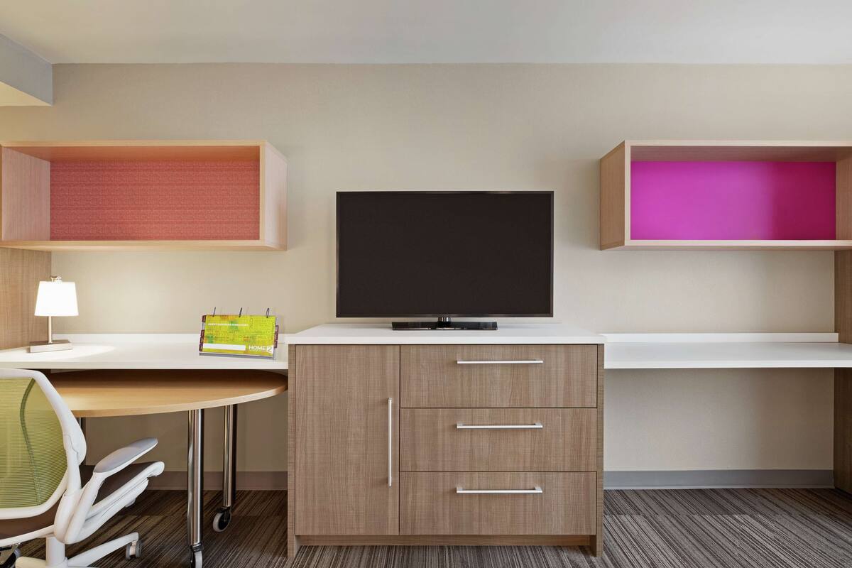 A hotel room with a desk, a television, a chair, and two shelves with storage compartments.