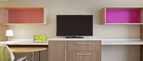 A hotel room with a desk, a television, a chair, and two shelves with storage compartments.