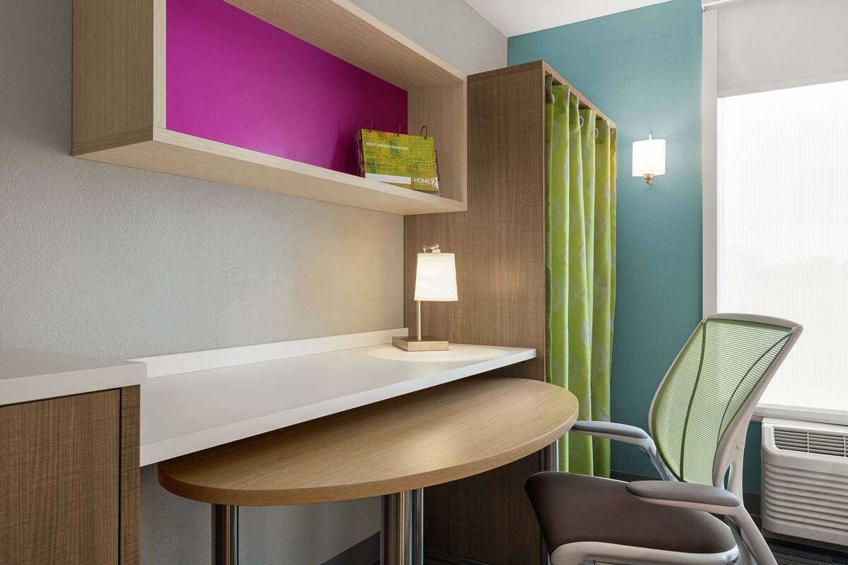 A modern room with a white desk, a green chair, a lamp, and a shelf with a colorful bag.