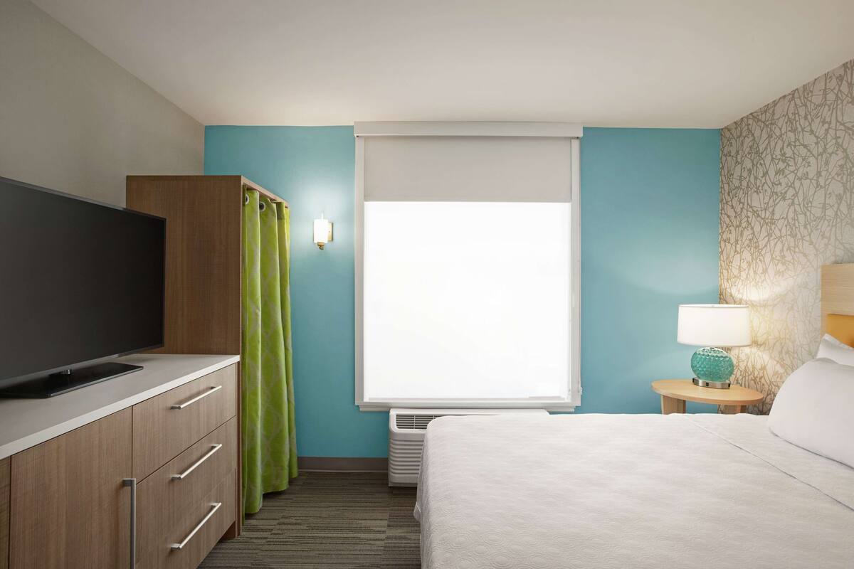 A hotel room with a bed, a TV, a wardrobe, a window with curtains, a lamp, and a small table.