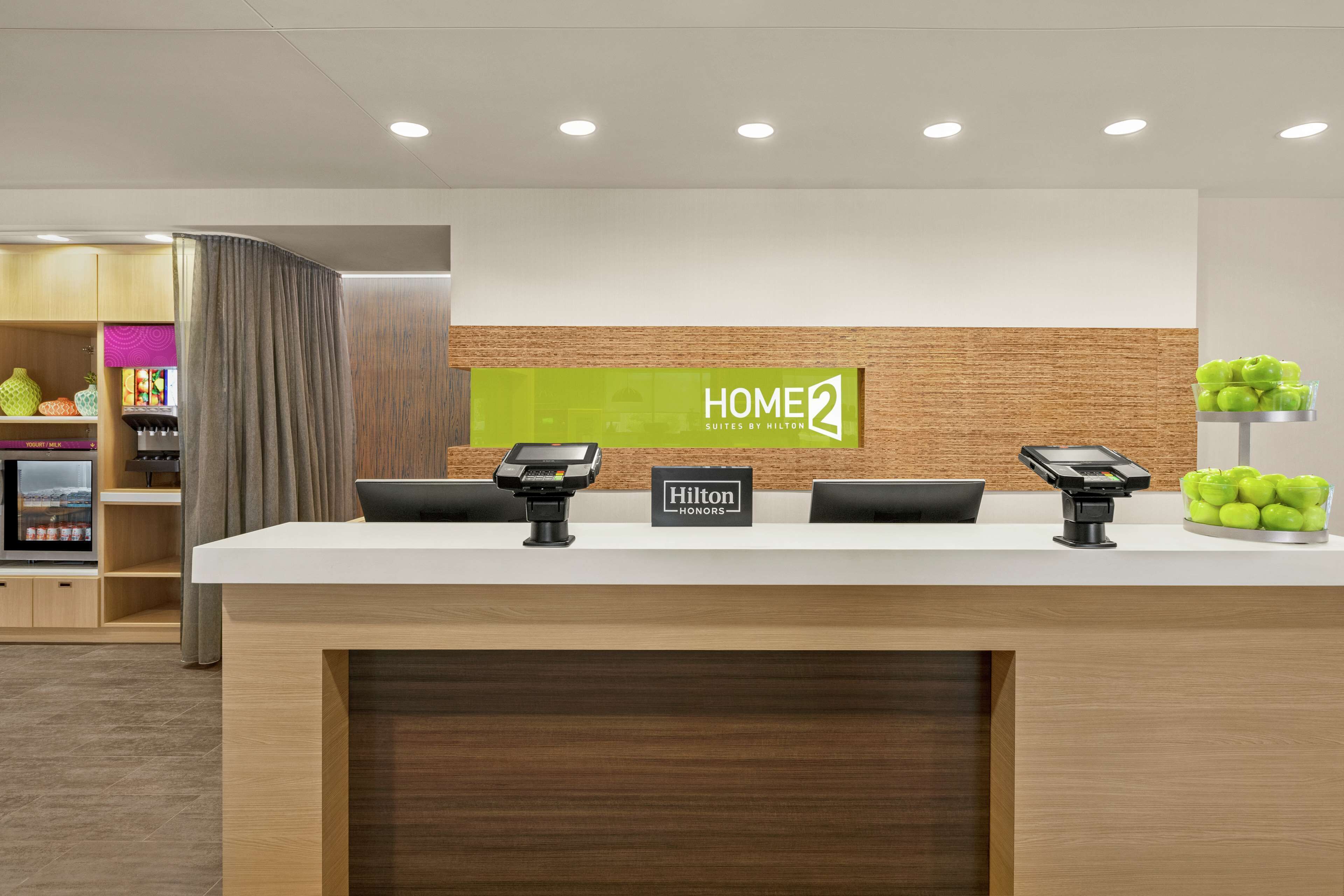 Un comptoir d’accueil d’hôtel avec une enseigne « HOME2 Suites by Hilton », un badge « Hilton Honors » et un kiosque numérique.