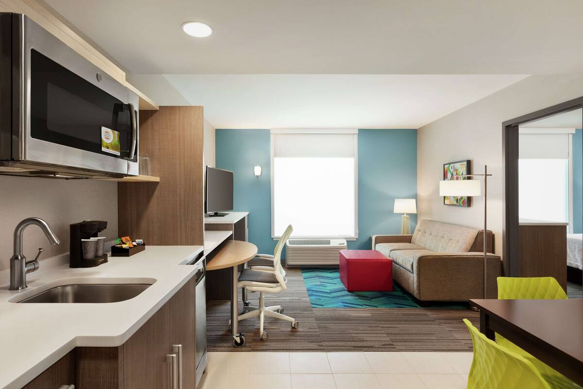 A modern hotel room with a kitchenette, a sofa, a dining table, and a large window providing natural light.