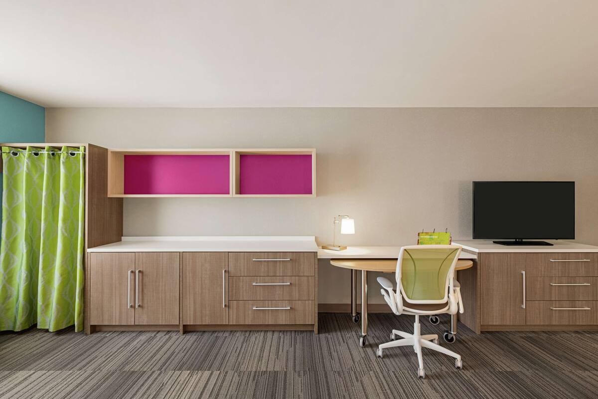 A hotel room with a desk, chair, TV, and two cabinets with pink shelves.