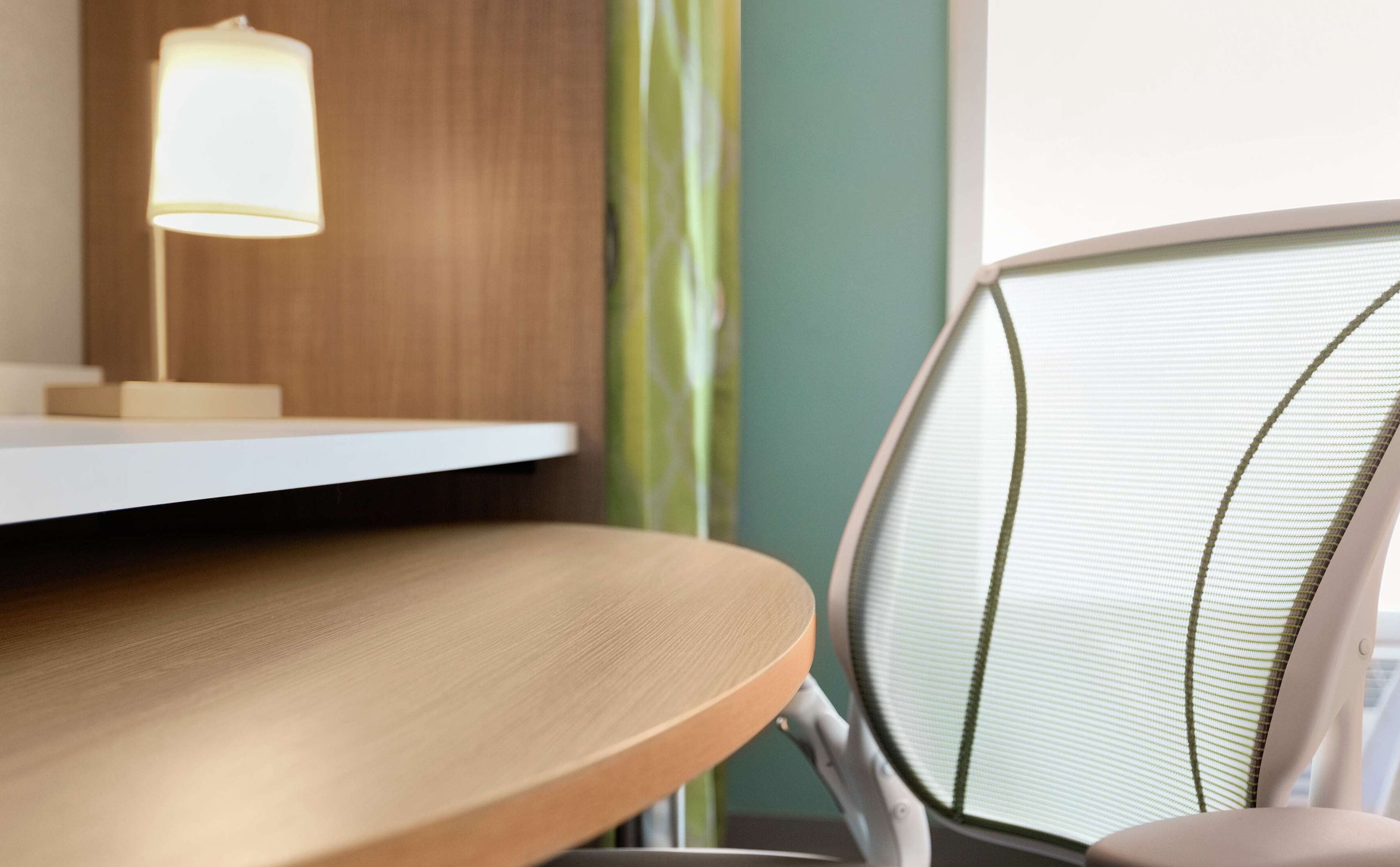 A wooden desk with a lamp, a chair with a mesh backrest, and a window with a green curtain.