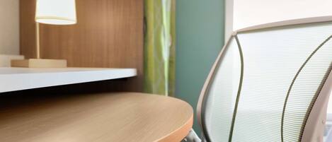 A wooden desk with a lamp, a chair with a mesh backrest, and a window with a green curtain.