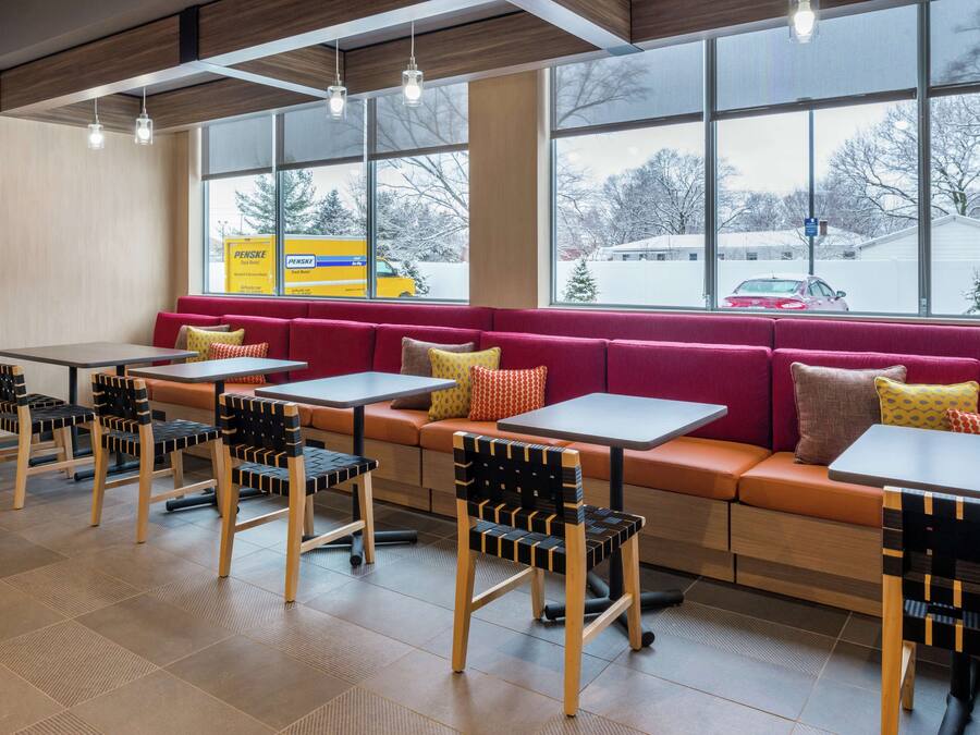 Un café moderno con mesas y sillas de madera, un banco con cojín rojo y grandes ventanales con vistas a un estacionamiento nevado.