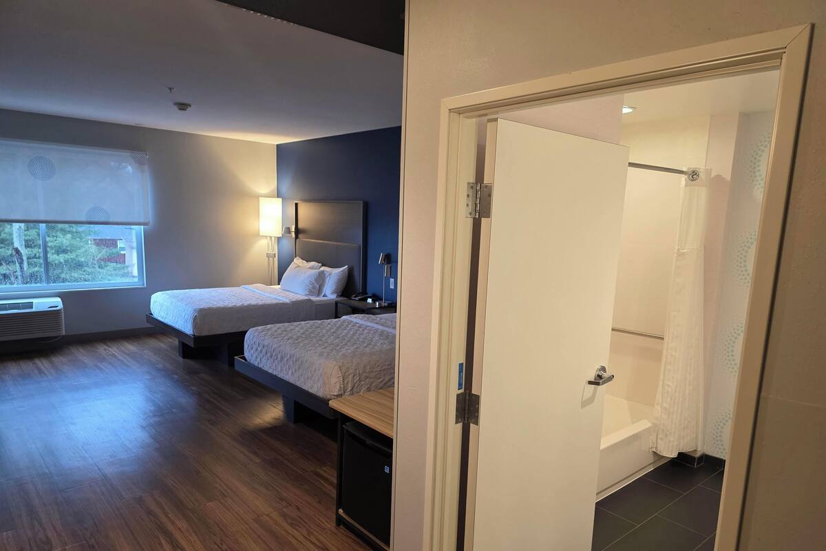 A hotel room with two beds, a window with blinds, a bedside lamp, a heater, and a door leading to a bathroom.