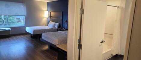Habitación de hotel con dos camas, una ventana con persianas, una lámpara de noche, un calentador y una puerta que da a un baño.