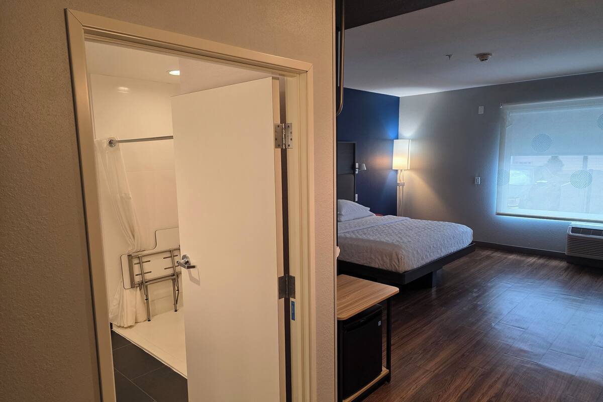 A modern hotel room with a wooden floor, a bed with a white duvet, a bedside table, a window with blinds, and a bathroom with a shower and a towel rack.