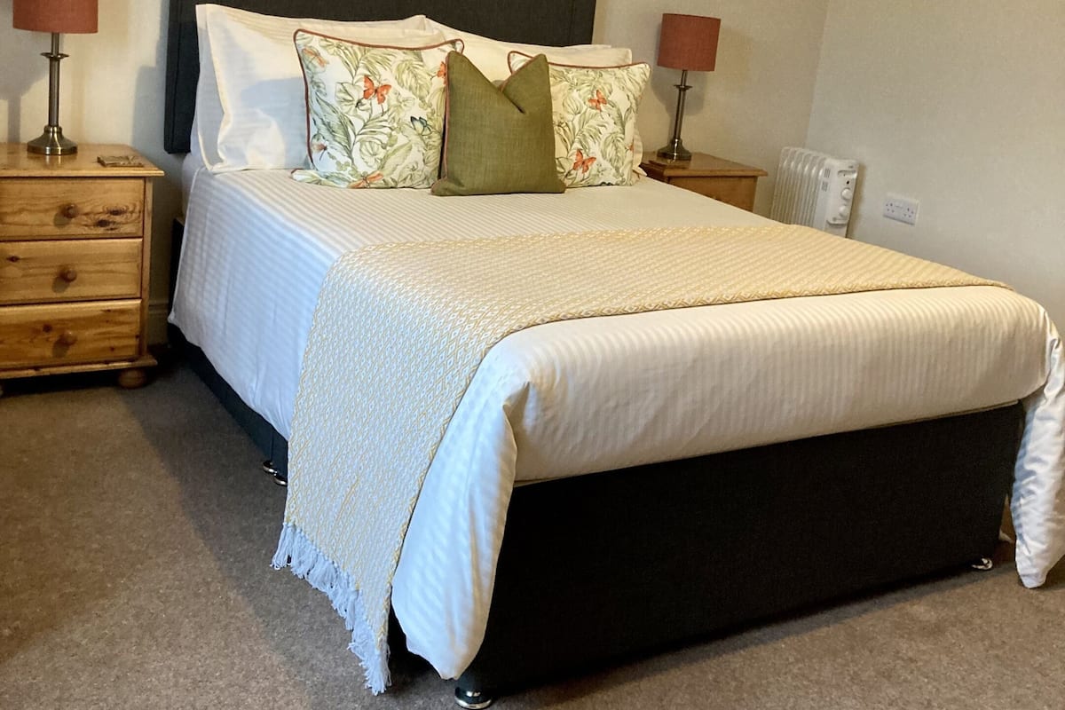 A neatly made bed with a dark headboard, a wooden nightstand with a lamp, and a radiator on the wall.