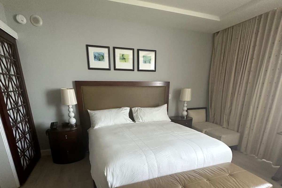 A hotel room with a large bed, a bedside table with a lamp, a small bedside table with a phone, a window with curtains, and a wall with three framed pictures.