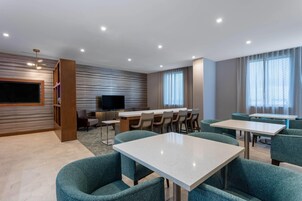 Una habitación de hotel moderna con un televisor de pantalla plana grande, una zona de comedor con mesas y sillas, y un amplio ventanal con cortinas.