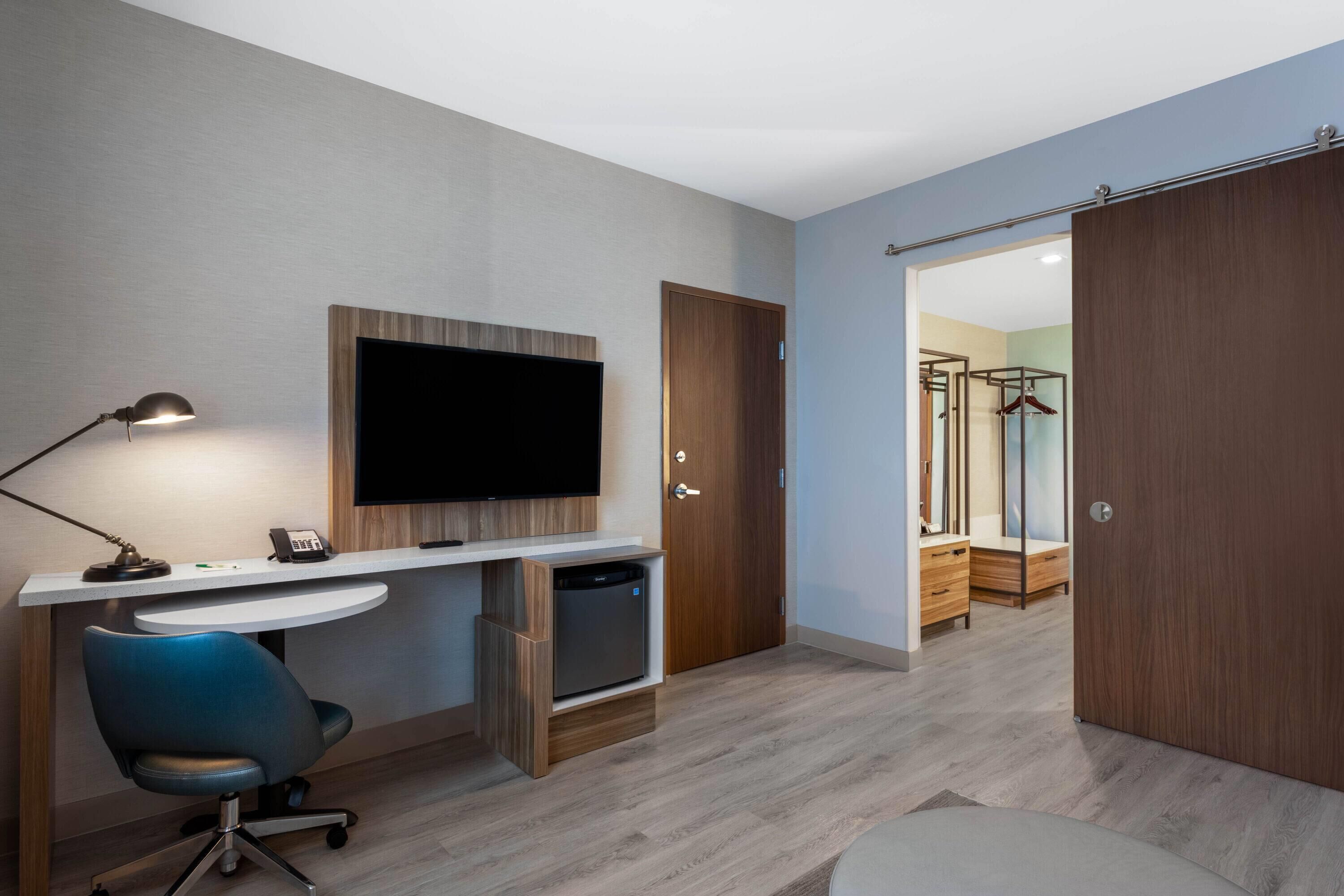 Una habitación de hotel moderna con escritorio, silla, televisor de pantalla plana, armario y vestidor.