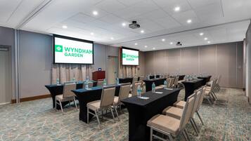 Un salón de conferencias con mesas y sillas, dos pantallas mostrando 'WYNDHAM GARDEN', y suelo alfombrado.