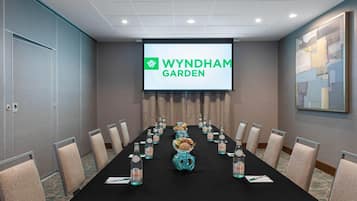 Un salón de conferencias con una larga mesa, sillas, una pantalla de proyector mostrando 'WYNDHAM GARDEN' y jarrones decorativos.