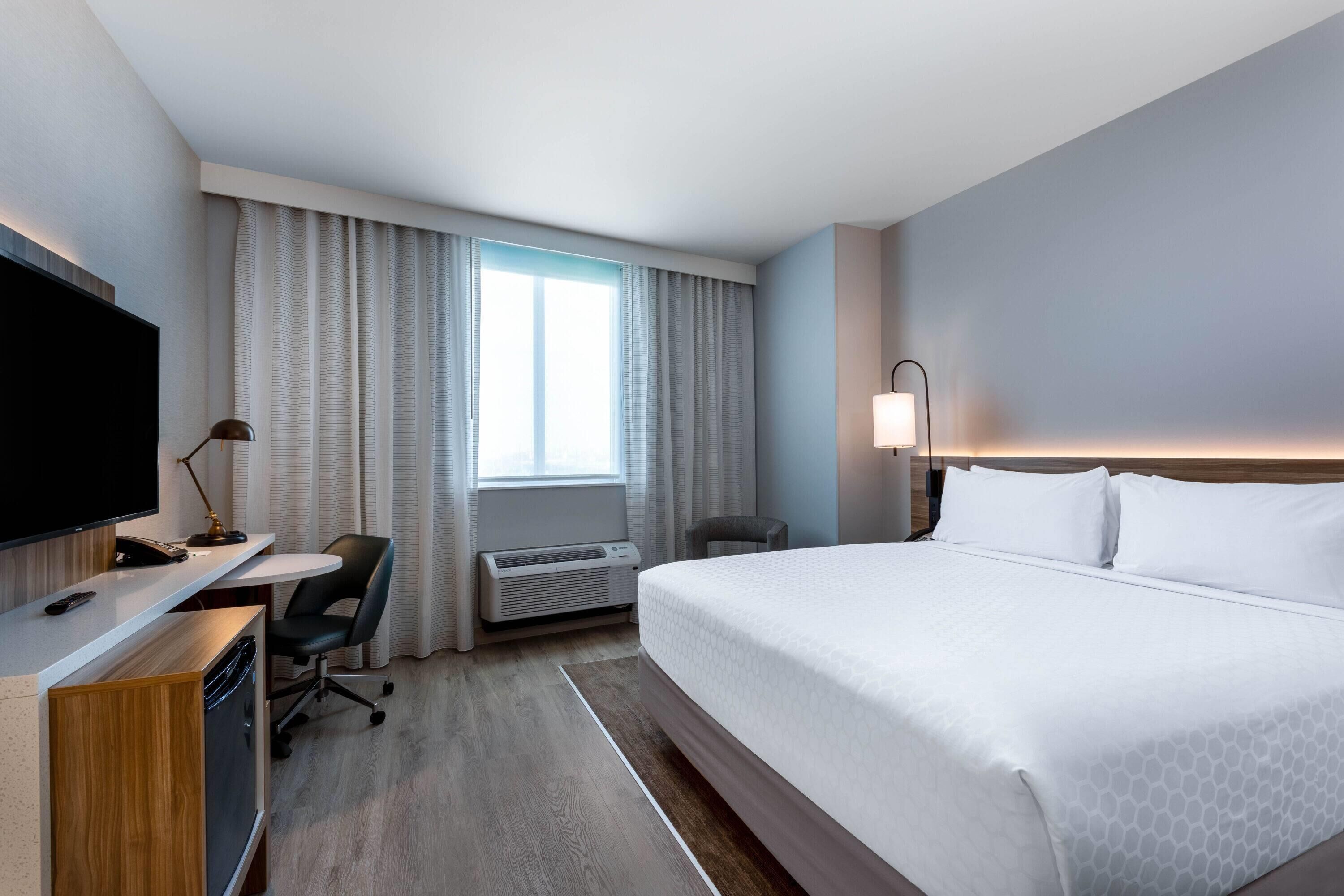 A hotel room with a bed, desk, chair, TV, air conditioning unit, and a window with curtains.